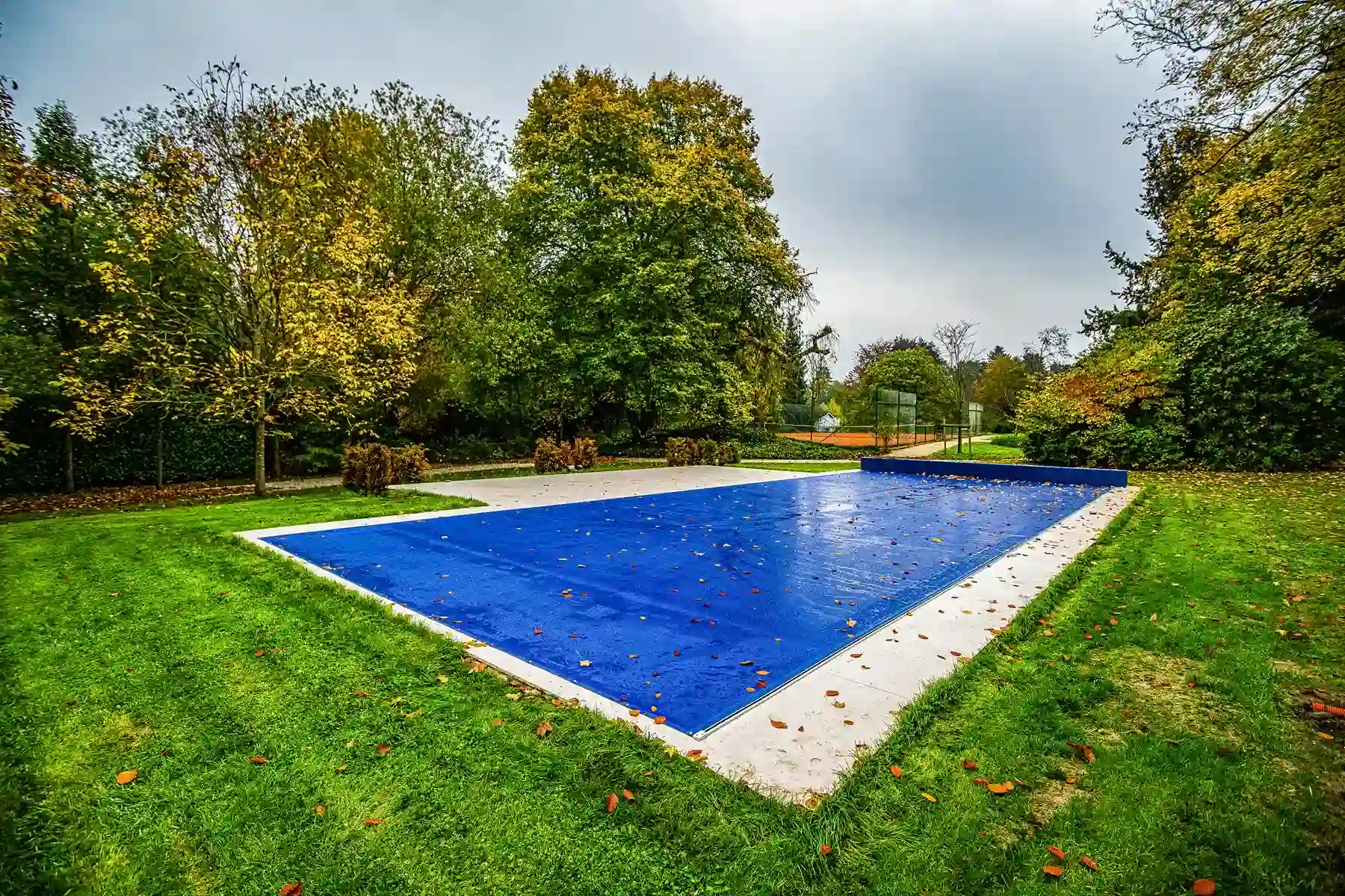 Piscine couverte d'une bâche bleue entourée de pelouse verte et d'arbres aux feuilles jaunissantes.