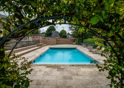 Piscine rectangulaire entourée de dalles en pierre, avec des chaises longues sur le côté droit et un arbre en arc au premier plan.