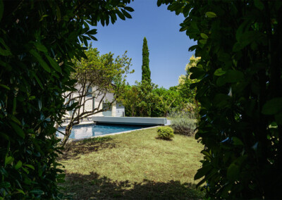 Sequoia4 Vue depuis le jardin encadrée par des feuillages, laissant apparaître la piscine et sa couverture Coverseal grise au cœur d’un écrin de verdure.