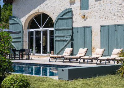 Sequoia5 Zoom sur la piscine protégée par une Coverseal grise, au pied d’une maison en pierre avec grande baie vitrée et volets verts.